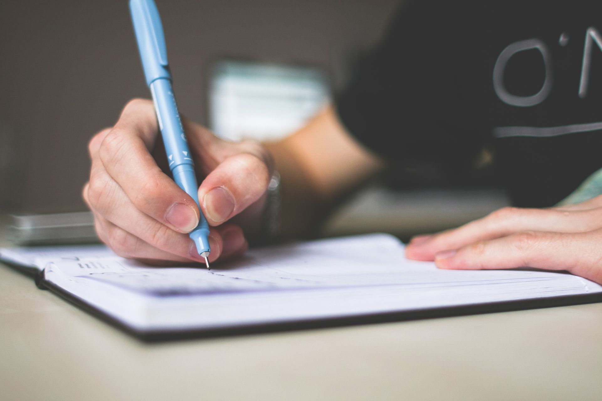 A hand writing in an open notebook with a pale blue pen at a warm wooden desk